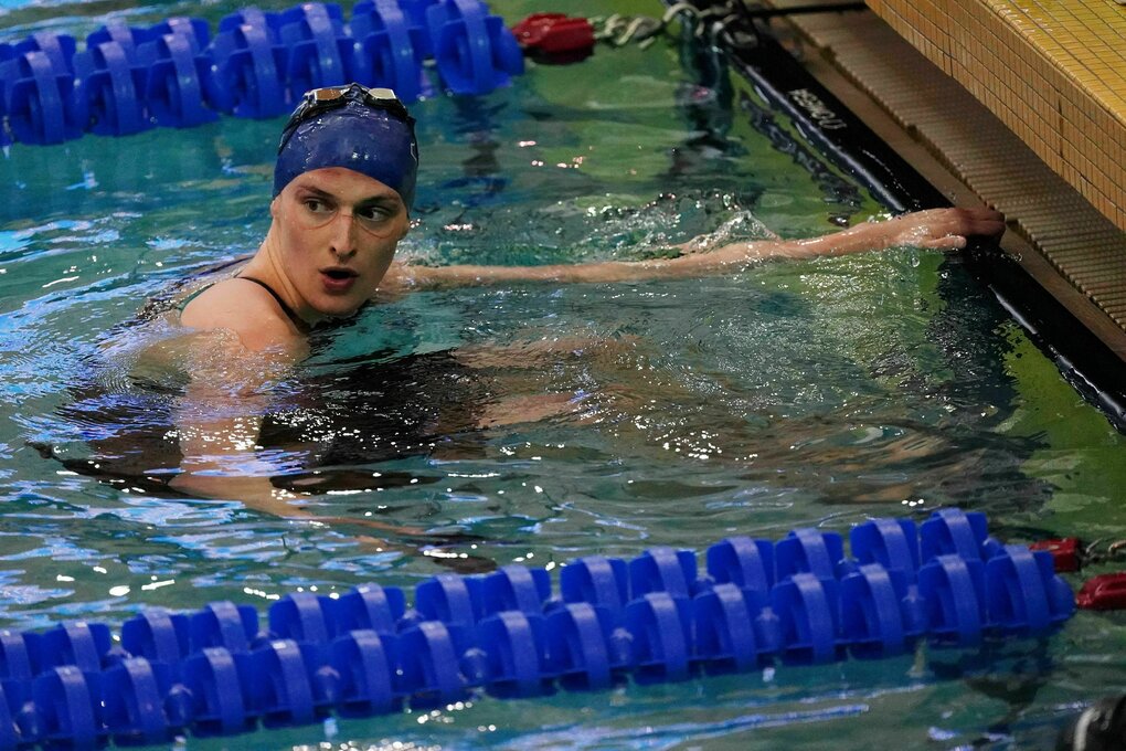 Lia Thomas im Schwimmbecken. Eine Junge Frau mit blauer Badekappe und Druckstellen rund um die Augen, wo die Schwimmbrille gesessen hat. Sie hält sich mit einer Hand am Beckenrand fest.