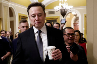 Elon Musk hält einen Kaffeebecher in der Hand. er ist ein mittelalter Mann mit kurzem, dunklem Haar. Er zieht die Mundwinkel nach unten. Hinter ihm drängen sich viele Menschen. Er ist in einem gelben Gang.