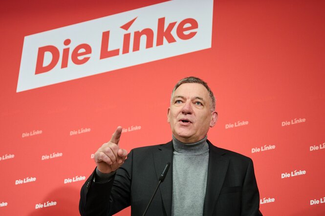 Jan van Aken spricht vor einer Pressewand der Linken und deutet bestimmt nach vorne