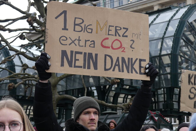 ein Mann hält ein Schild hoch, auf dem steht: "1 Big Merz mit extra CO2 - NEIN DANKE!!