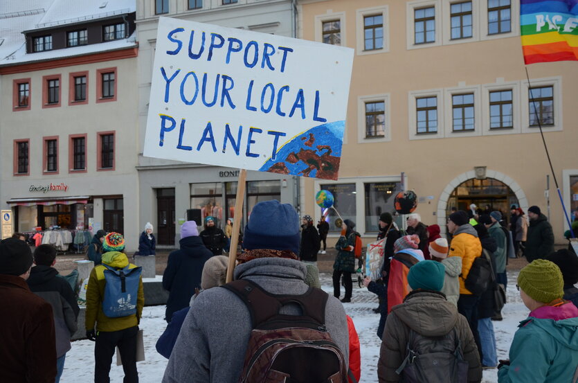 Mehrere Menschen auf einem winterlichen Stadtplatz, einer hält ein Plakat hoch auf dem steht: Support your local planet