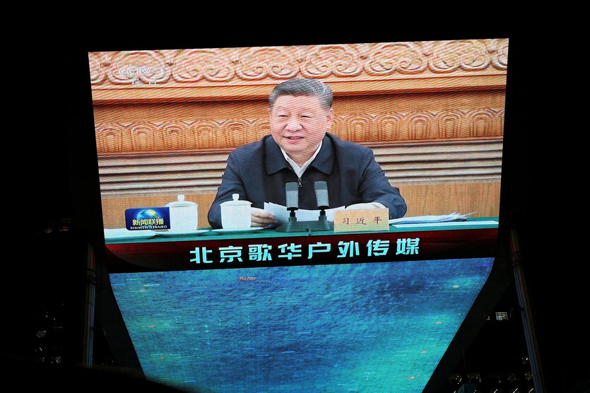 Xi Jinping ist auf einer Leinwand zu sehen, er sitzt an einem Tisch, vor ihm Mikrofone und zwei Teetassen