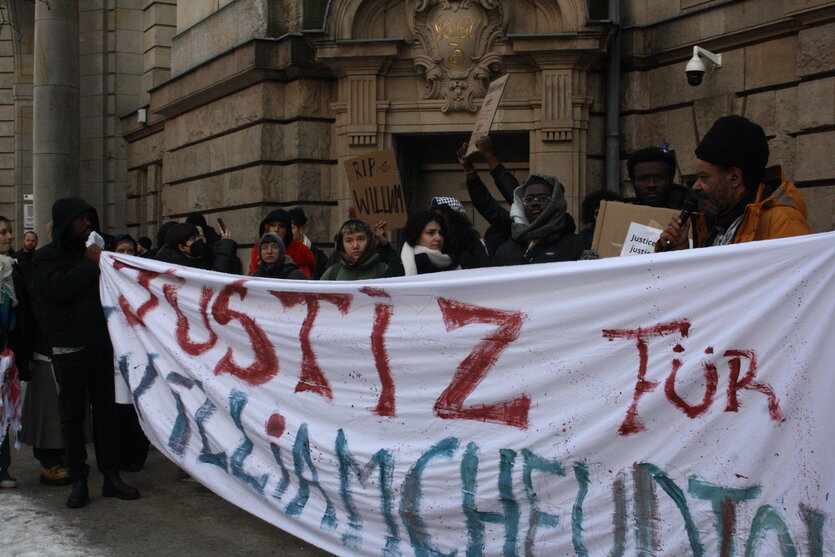 Menschen stehen vor dem Berliner Strafgericht in Moabit und halten Transparente in Solidarität mit William Chedjou hoch