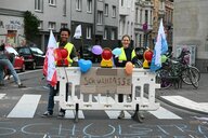 Zwei Personen stehen lächelnd hinter einer Straßenabsperrung an der ein Schild hängt, auf dem Schulstraße steht