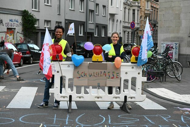 Zwei Personen stehen lächelnd hinter einer Straßenabsperrung an der ein Schild hängt, auf dem Schulstraße steht