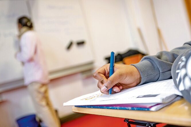 Eine Hand schreibt mit Füller in ein Heft, dahinter steht eine Lehrerin