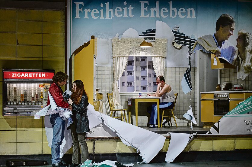 Ein junges Paar steht vor einem halb herunter gerissenen Werbeplakat. Die Löcher gegen den Blick frei in eine triste Wohnung in der ein Mann an einem Tisch sitzt.