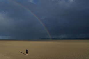 Eine Person steht auf einem menschenleeren Sandstrand und schaut auf einen Regenbogen, der Hmmel ist dunkelblau