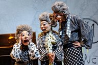 Drei mit Masken und Damenkleidung kostümierte Schauspieler und Schauspielerinnen