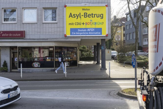 Ein gelbes Plakat hängt in einer grauen Innenstadt: Drauf steht "Weiter Asyl-Betrug mit CDU + Rot/Grün? Deshalb AFD Die bürgerliche Alternative"