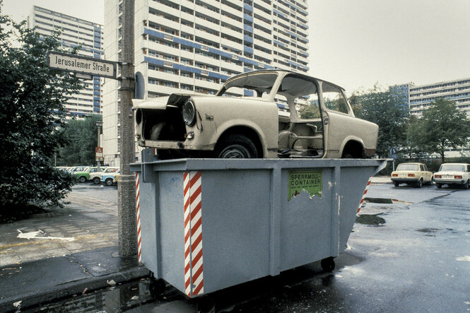 Ein Trabbi ohne Räder, etwas derangiert, steckt obenauf in einem Müllcontainer