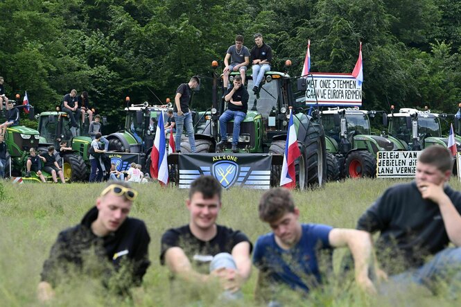 Bauernprotest in Brüssel - junge Bauern stehen mit ihren großen Traktoren auf einer Weise - vier junge Männer sitzen im Gras