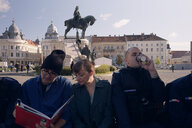 Eine Gruppe Menschen sitzt auf einer Bank an einem zentralen Platz in Cluj, im Hintergrund ein Reiterdenkmal und die barocke Silhouette der rumänischen Stadt, einer der Personen ist ein Polizist und trinkt gerade aus einem ToGo-Becher