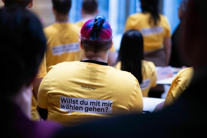 Ansicht auf einen Rücken in gelbem Tshit, mit der Aufschrift "Willst du mit mir wählen gehen?", die Person hat lila-blaue Haare in einem Dutt