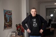 Jan van Aken posiert in einem Wahlkreisbüro. Er trägt ein T-shirt mit der Aufschrift " Tax the Rich"