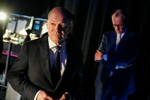 Olaf Scholz (l) und Friedrich Merz (r) warten auf den Beginn des "Quadrell"; Scholz blick besorgt in die Ferne, Merz blickt betroffen zu Boden