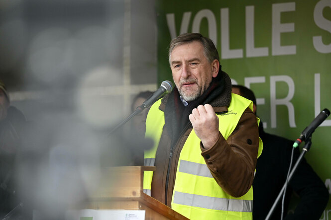 Ein Mann mit grau-melliertem Bart und Haupthaar und verhärteten Gesichtszügen in Warnweste hebt die Faust geballt drohend auf einer Bühne.