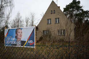 Ein Wahlplakat zur Bundestagswahl 2025 mit der AfD-Bundesvorsitzende, Alice Weidel, steht in einem tristen Vorgarten mit einem unbewohnten Haus.