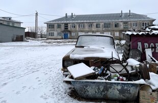 Vor einem Plattenbau stehen auf einem schneebedeckten Platz eine Badewanne voller schrott und ein altes Auto