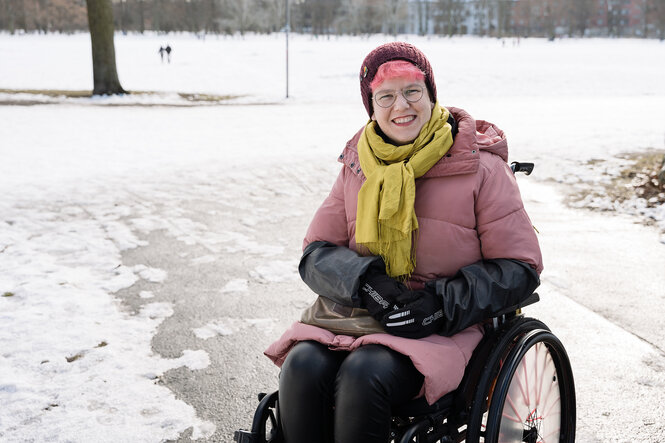 Eine Frau sitzt im Rollstuhl, trägt rosa gefärbte Haare und eine rosa Winterjacke