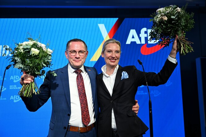 Tino Chrupalla und Alice Weidel mit Blumen vor einem blauen AfD-Hintergrund bei einer Wahlkampfparty