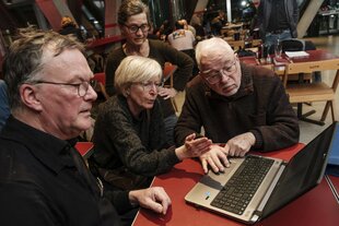 Zu sehen ist: Drei Menschen sitzen in der taz Kantine gemeinsam an einem Tisch und blicken in einen Laptop. Zwei der Menschen befinden sich womöglich im Rentenalter.