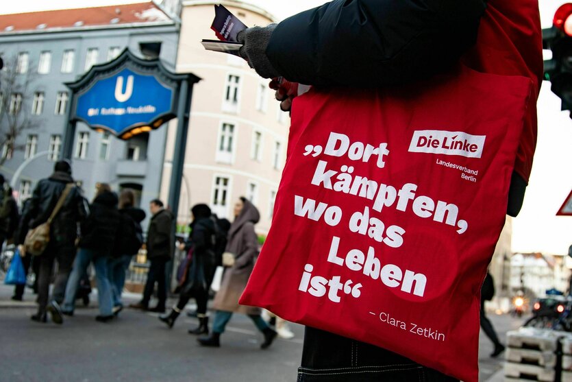 Straße in Neukölln, Blick auf den U-Bahn eingang, Menschen unterwegs auf der Straße. Eine Person verteilt Flyer und die rote Stofftasche leuchtet, sie ist mit einem Clara Zetkin Zitat beschriftet: Dort kämpfen, wo das Leben ist