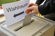 Das Bild zeigt eine Hand, die einen Wahlsein in den Schlitz einer Wahlurne steckt