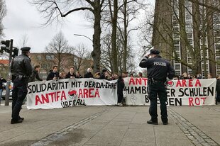 Zwei Polizisten vor zwei Transparenten, dahinter weitere Menschen