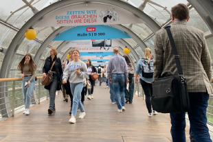 Zu sehen ist: Ein lichtdurchfluteter Glasgang der Messe Leipzig, der von Besucher*innen gefüllt ist.