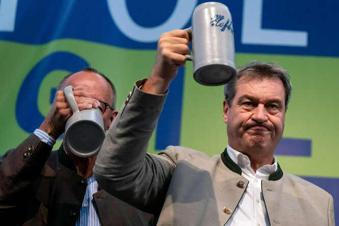 Markus Söder hebt einen Steinhumpen prostend in die Höhe, Merz hinter ihm hat den Kopf bereits in einem der selbigen versenkt