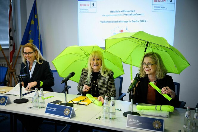 Drei Frauen sitzen lachend auf einem Podium, zwei davon haben über sich einen neongelben Regenschirm aufgespannt