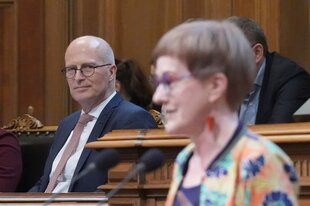 Eine Frau mit Brille in bunter Jacke, dahinter sitze ein Mann im Anzug