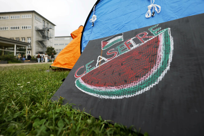 Vordergrund; ein gemaltes Schild mit einer Wassermelone, kleiner Palestina Flagge und "Ceasefire" Aufschrift, an einem Zelt lehnend. Im Hintergrund ein Uni Gebäude