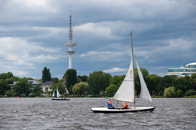 Ein Segelboot auf grauem Wasser, dahinter Bäume und Fernsehturm