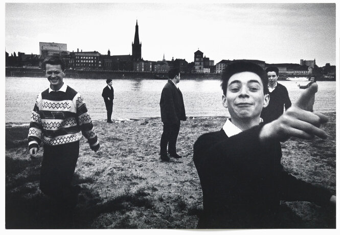 Schwarz-Weiß-Fotografie von Leonard Freed: Ein Gruppe Jugendlicher spielt am Flussufer in Düsseldorf. Ein Junge läuft auf die Kamera zu und zeigt lächelnd hinter die Linse.