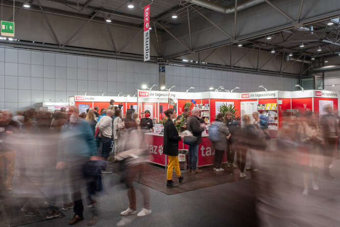 Das Bild zeigt den taz Stand auf der Leipziger Buchmesse. Es sind Menschen verschwommen zu sehen, in der Mitte ist eine taz Mitarbeiterin zu sehen, die Zeitungen verteilt