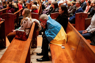 Ein Mann sitzt auf einer Kirchenbank, er hat eine Fahne in den Nationalfarben der Ukraine um die Schultern gelegt