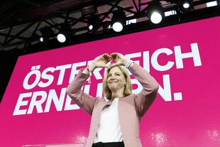 Eine Frau macht das Herzchen-Zeichen vor einer pinken Wand mit der Aufschrift