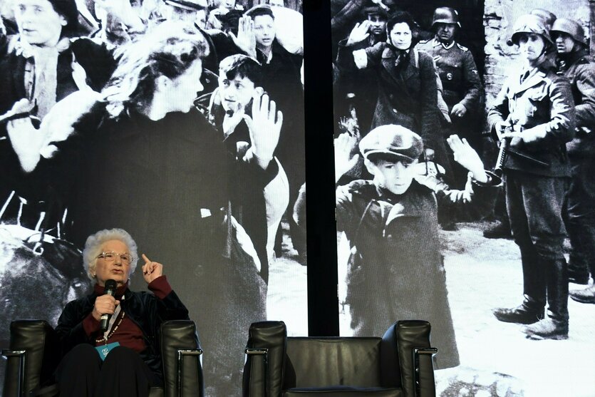 Eine Frau sitzt auf einer Bühne und spricht mit erhobenen Zeigefinger in ein Mikrofon. Hinter ihr ist ein großes Foto zu sehen, auf dem SS-Truppen Bewohner des Warschauer Ghettos deportieren