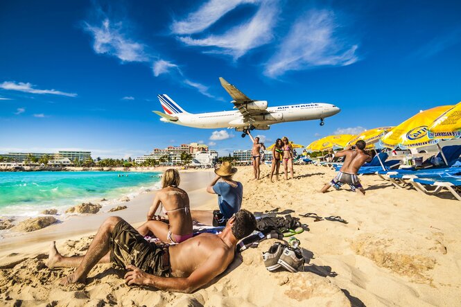 Eine Airfrance Maschine landet tief, Menschen liegen am STrand im Sand und schauen auf das Flugzeug, im Hintergrund Bettenburgen