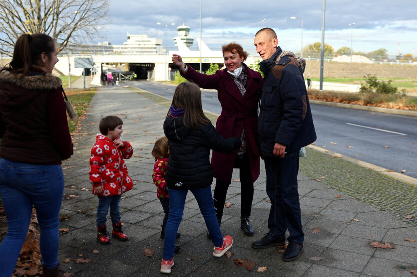 Ein Mann und eine Frau begrüssen drei Kinder und eine Frau, im Hintergrund der ehemalige Flughafen Berlin