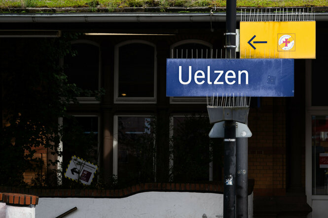 Foto zeigt das Bahnhofsschild mit der Aufschrift "Uelzen", dahinter sind dunkel und schemenhaft Teile des Bahnhofs zu erkennen.