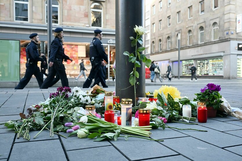 An einem Laternenpfahl liegen Blumen und Grablichter auf dem Asphalt, im Hintergrund geht eine Gruppe von Polizisten