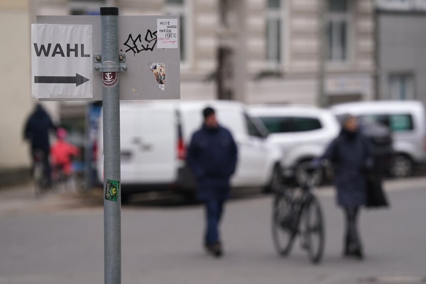 Im Vordergrund ein Straßenschild mit einem Zettel mit der Aufschrift "Wahl" und einem Pfeil. Im Hintergrund laufen Menschen auf der Straße in Richtung des Pfeils.