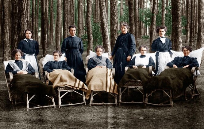 Frauen in der Tracht des frühen 20. Jahrhunderts in Liegestühlen, die in einem Wald stehen. Dahinter Pflegerinnen.