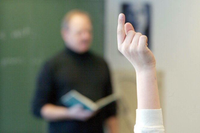 Eine Person meldet sich während einer Schulstunde. Der Lehrer, mit einem Buch in der Hand, ist unscharf im Hintergrund zu sehen