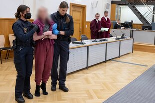 Eine Frau in roter Kleidung steht auf Zehenspitzen und Socken im Gerichtssaal, sie wird von 2 Justizbeamtinnen festgehalten