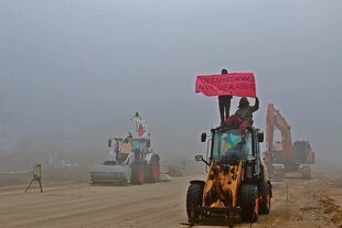 Klimaaktivist:innen besetzen einen Bagger, eine Walze und einen Radlader auf einer Baustelle und halten Transparente hoch, Aufschrift: Verkehrswende? Nur sozialgerecht!!!