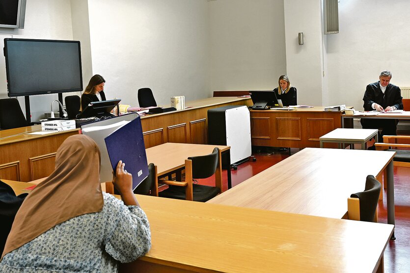 Ein Gerichtssaal im Strafjustizgebäude in Hamburg. Eine Frau mit Kopftuch ist von hinten zu sehen, sie hält sich einen Ordner vor das Gesicht. Im Hintergrund sieht man die Richterin und den Nebenklagevertreter.